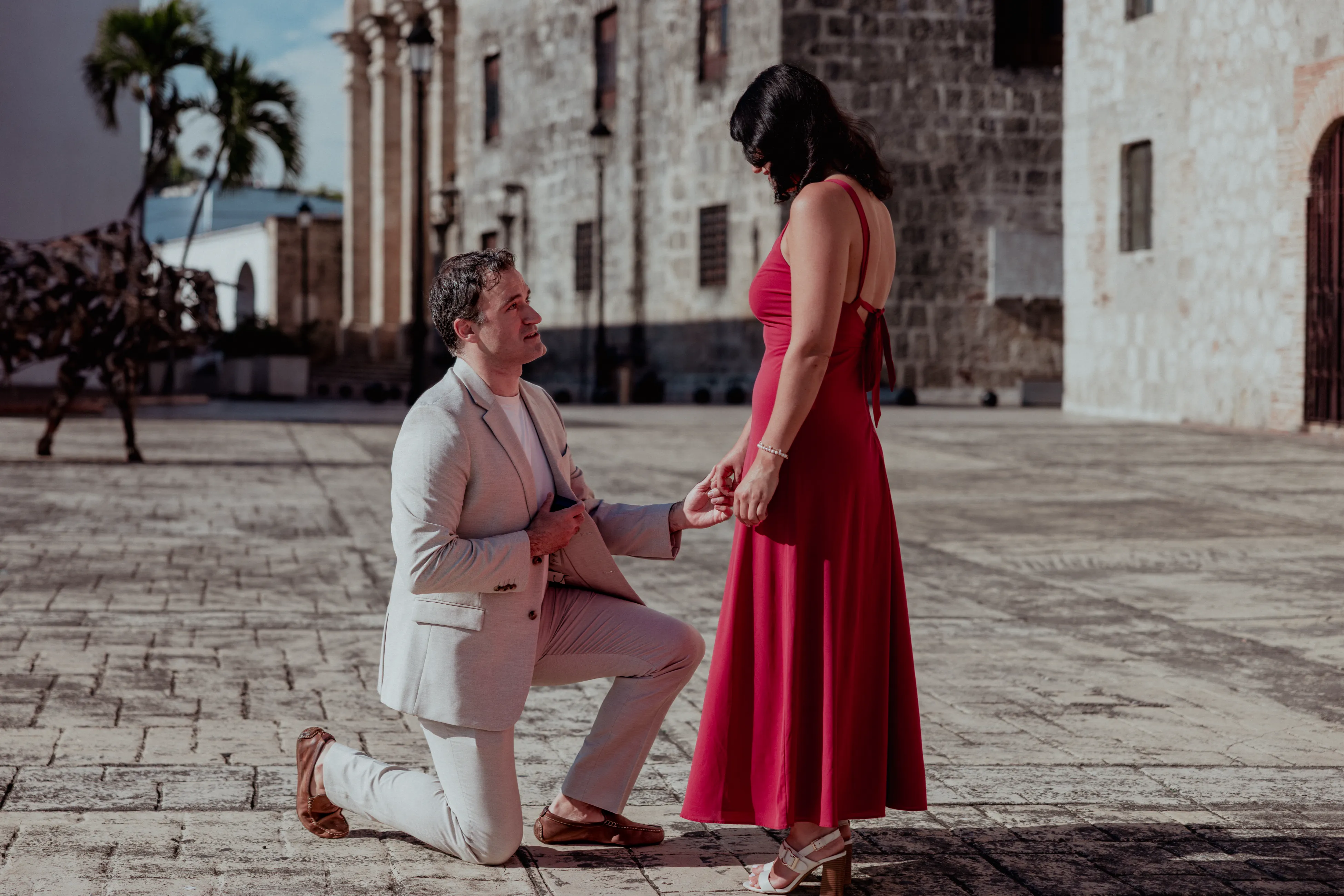 Proposal in the Colonial Zone Man in white suit kneeling before woman in red dress in Santo Domingo colonial plaza proposal photography moment