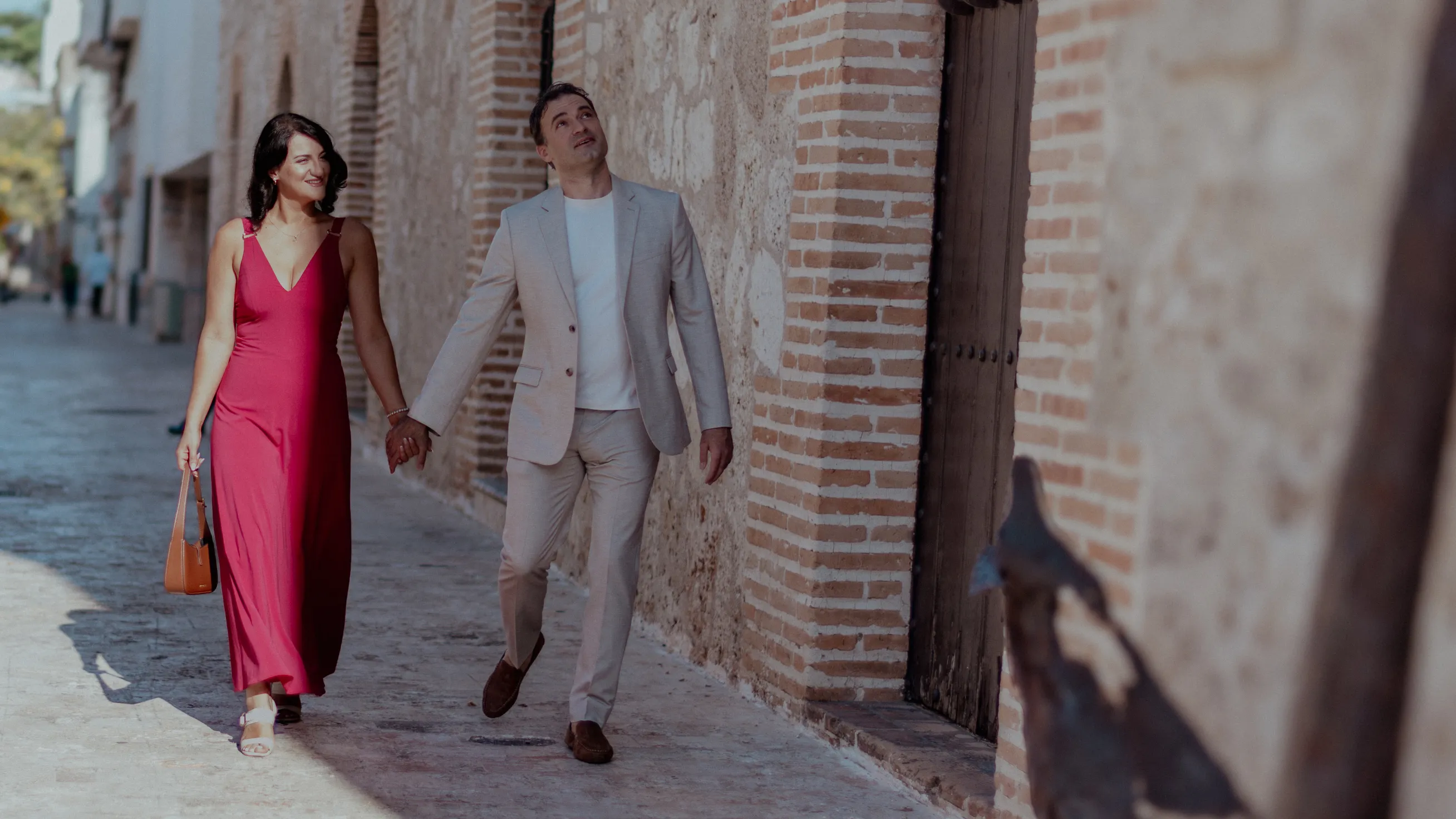 Love in motion through the Colonial Zone Couple walking on cobblestone streets of Santo Domingo's Colonial Zone; she wears deep red dress, he wears beige suit. Natural midday light