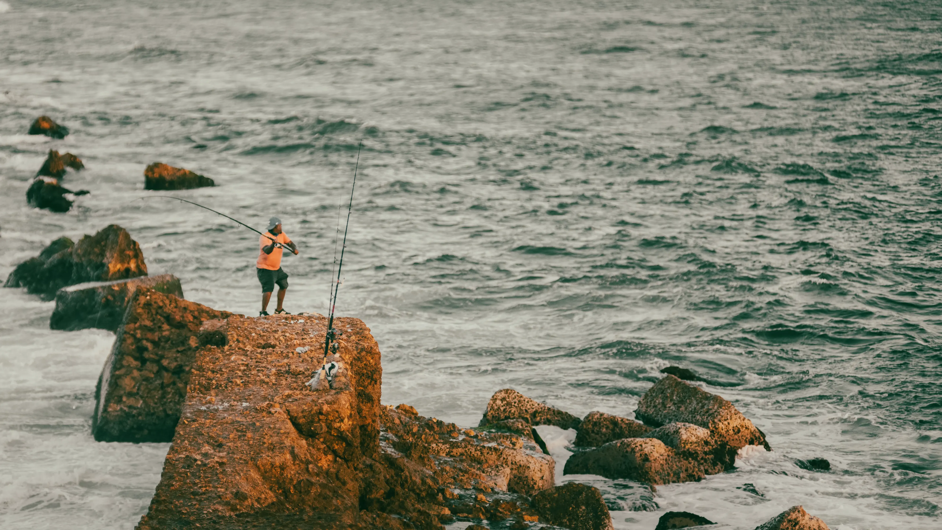 Pescador en la costa dominicana Pescador en chaqueta naranja parado sobre rocas oxidadas lanzando caña de pescar hacia el mar en costa dominicana