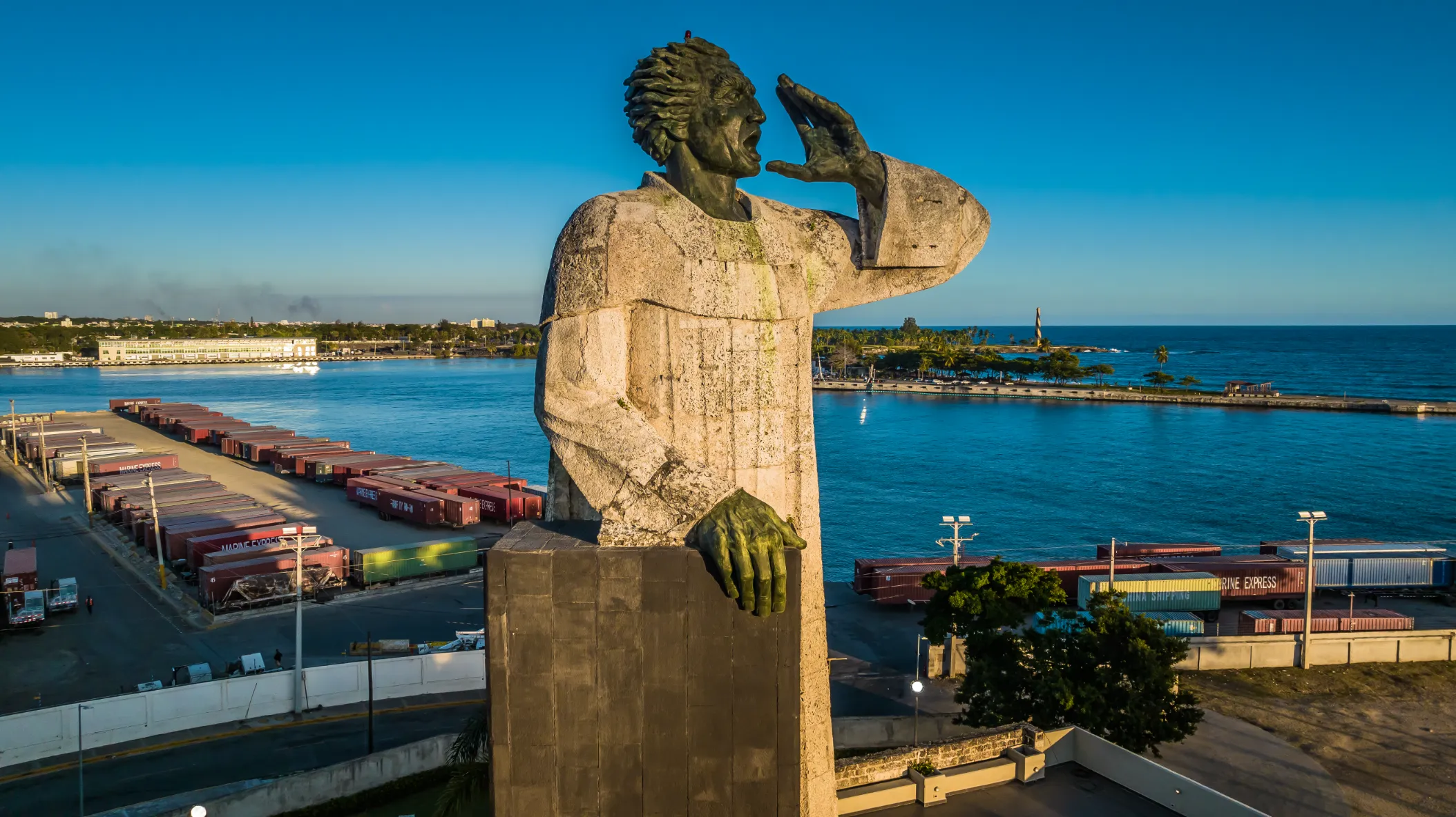 Harbor monument in golden morning light Golden stone statue with hand to cheek overlooking Santo Domingo port with containers and turquoise water, commercial photography