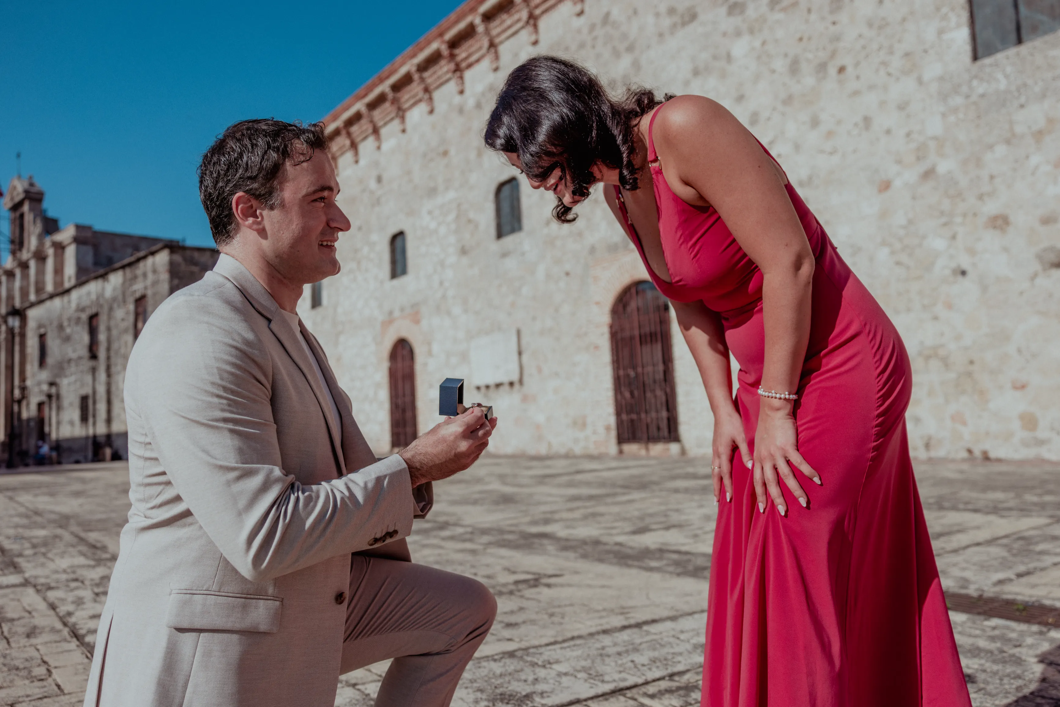 Proposal Under Santo Domingo's Blue Sky Man in beige suit proposes with diamond ring to woman in fuchsia dress in Santo Domingo's colonial plaza