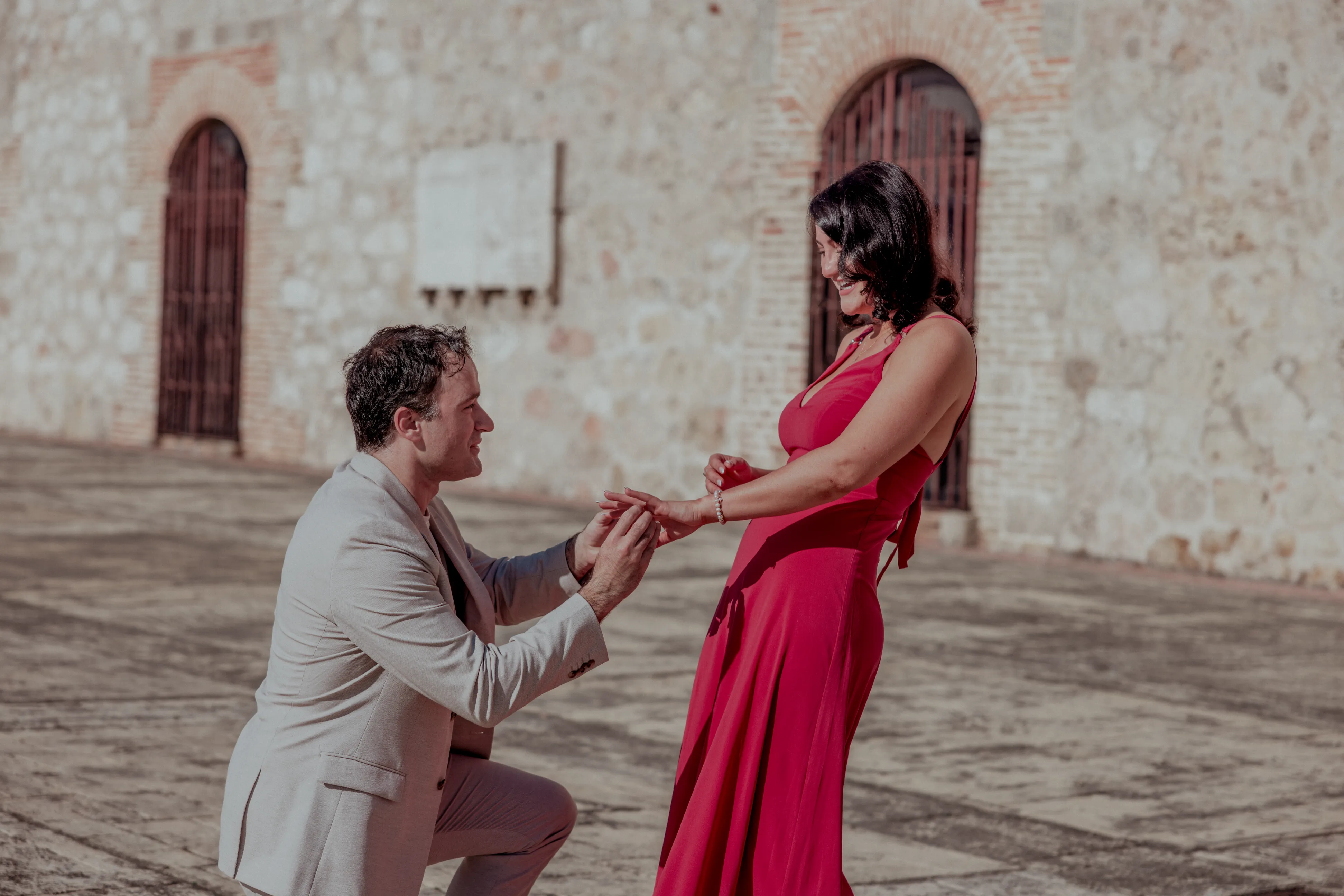 Marriage proposal in Colonial Zone Man in beige suit kneels with ring before woman in fuchsia dress in colonial Santo Domingo plaza