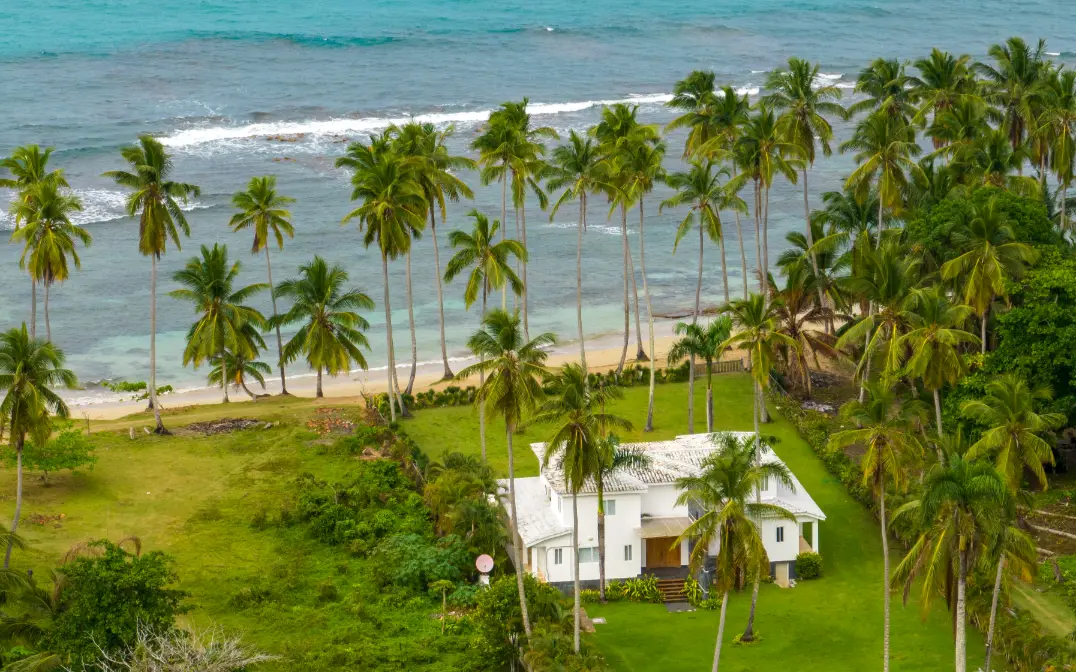 Playa con palmeras y casa costera Casa blanca rodeada de palmeras altas frente a playa turquesa con olas, sesión de familia playa República Dominicana